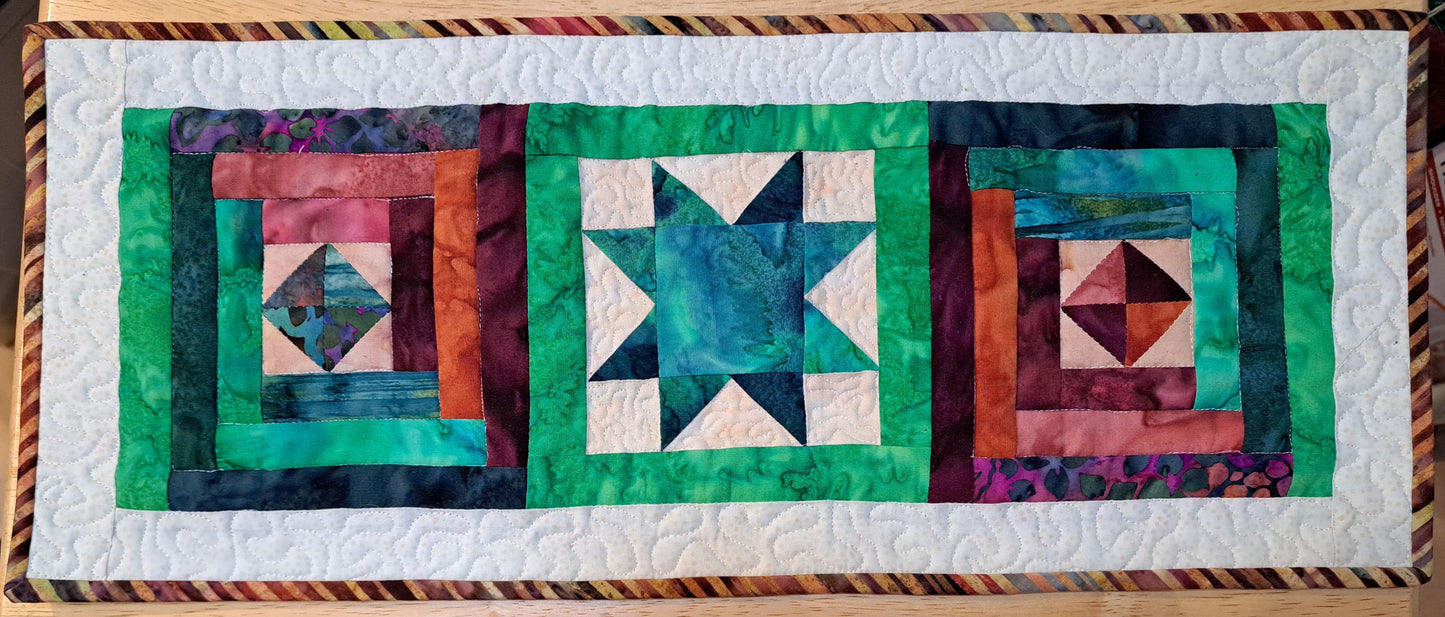 A quilted table topper/trivet with green, red, and beige geometric patterns and a striped batik binding.